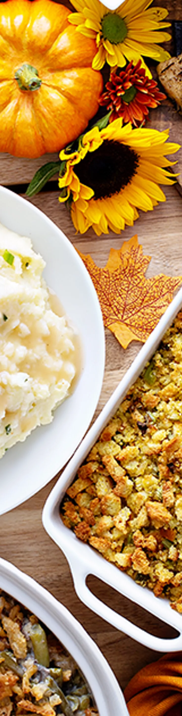 A festive spread of classic Thanksgiving dishes is arranged on a table, featuring sliced turkey, glazed ham, mashed potatoes, stuffing, and roasted carrots. The scene includes pumpkin pie with whipped cream, green bean casserole, and cornbread, surrounded by autumn decorations like pumpkins and leaves. Warm, golden tones and a cozy atmosphere evoke a celebratory holiday meal.