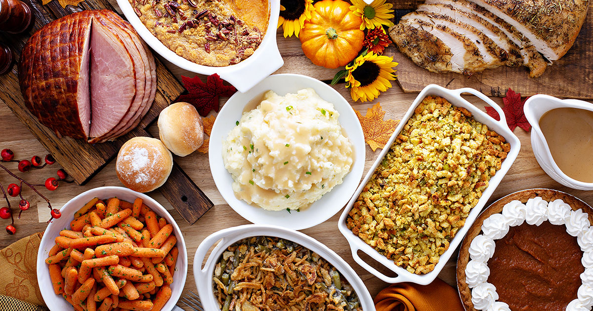 A festive spread of classic Thanksgiving dishes is arranged on a table, featuring sliced turkey, glazed ham, mashed potatoes, stuffing, and roasted carrots. The scene includes pumpkin pie with whipped cream, green bean casserole, and cornbread, surrounded by autumn decorations like pumpkins and leaves. Warm, golden tones and a cozy atmosphere evoke a celebratory holiday meal.