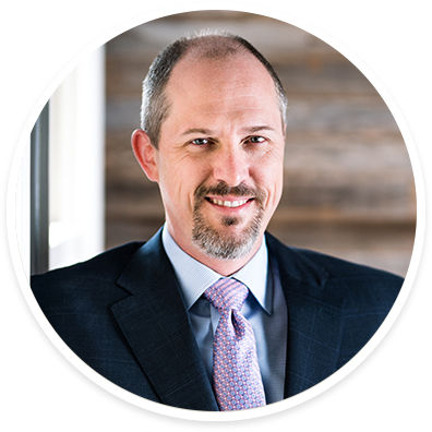 A person dressed in a dark suit, light blue shirt, and patterned tie is seated indoors. The background features wooden paneling and a window, suggesting a modern office or professional setting. The overall mood is formal and businesslike, with neutral and muted tones. The subject appears to be an adult male.