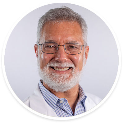 A mature male with gray hair is shown wearing a white lab coat over a blue striped shirt. The subject is photographed against a plain, light background, suggesting a professional or clinical setting. The image conveys a sense of professionalism and expertise, with a neutral and clean visual style. No visible text or numbers are present in the image.