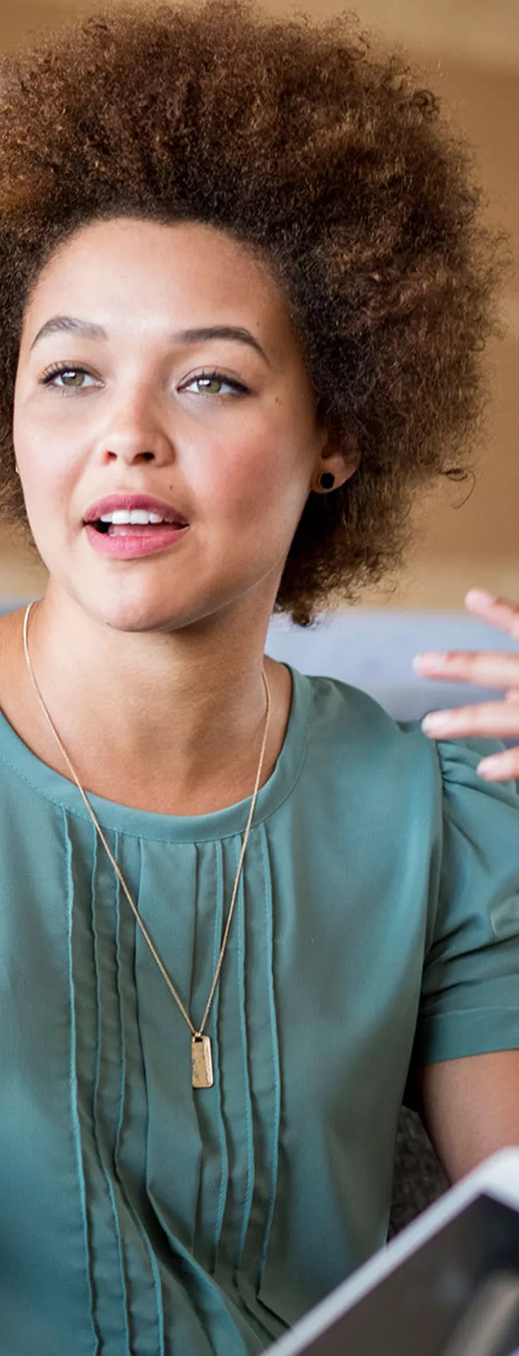 Two women are engaged in a discussion while seated on comfortable couches in a contemporary lounge or office space. The setting features neutral tones, framed wall art, and soft furnishings, creating a relaxed and professional atmosphere. One woman gestures expressively while the other listens, holding a tablet device. The overall mood is collaborative and informal.