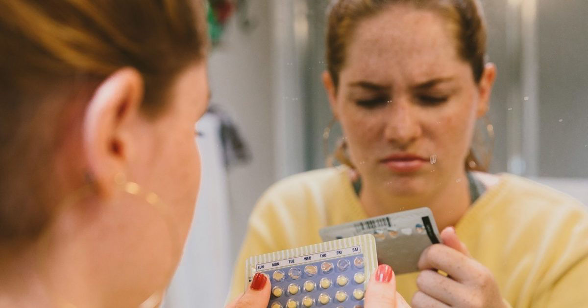 A woman stands in front of a bathroom mirror, holding a blister pack of birth control pills and a card. The setting is a home bathroom, with soft lighting and a casual atmosphere. The focus is on the hand holding the pill pack, with visible details of the pills. The image conveys a routine or daily health-related activity.