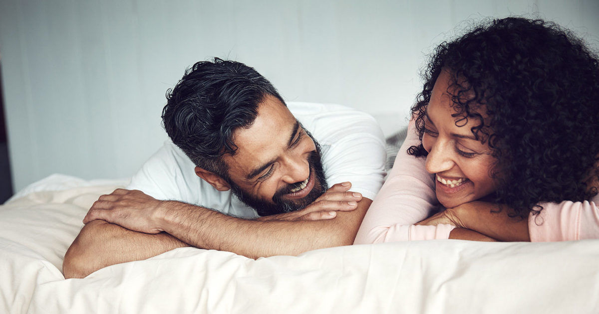 Two adults are lying on a bed, facing each other in a calm and intimate home environment. The bedding is light-colored, and the background is softly lit, creating a peaceful atmosphere. Both individuals appear to be engaged in a quiet moment, suggesting comfort and relaxation. No visible text or numeric values are present in the image.