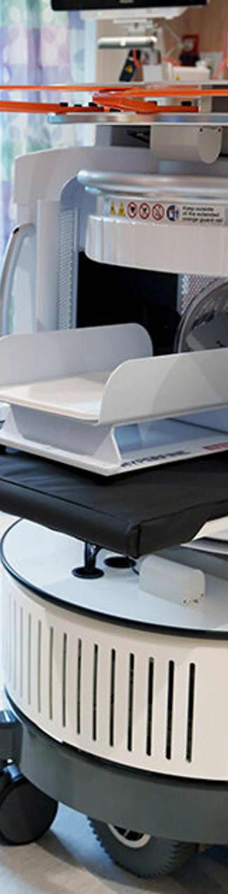 A hospital staff member stands in a modern medical room, interacting with a large autonomous robot labeled 'HYPER.' The robot features a tray system and a prominent orange rotor, suggesting possible drone or automated delivery capabilities. The staff member holds a tablet, indicating digital control or monitoring. The setting is clean and clinical, with wood paneling and medical equipment visible.