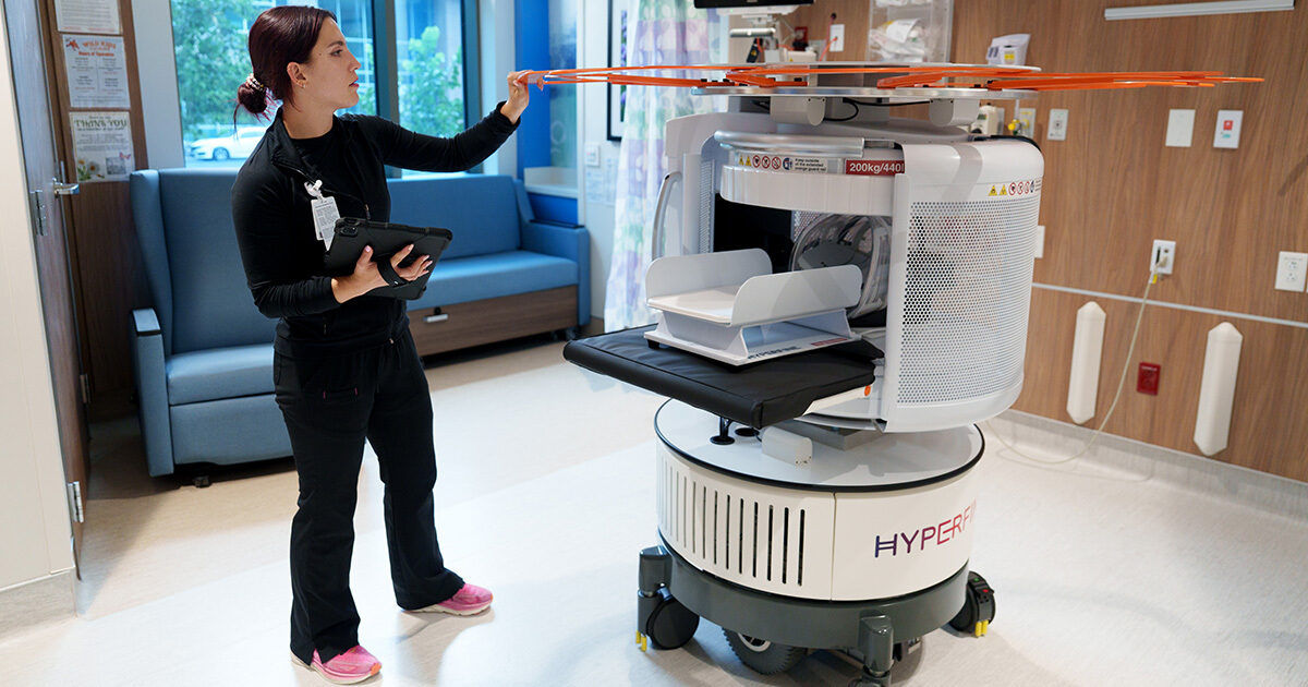A hospital staff member stands in a modern medical room, interacting with a large autonomous robot labeled 'HYPER.' The robot features a tray system and a prominent orange rotor, suggesting possible drone or automated delivery capabilities. The staff member holds a tablet, indicating digital control or monitoring. The setting is clean and clinical, with wood paneling and medical equipment visible.