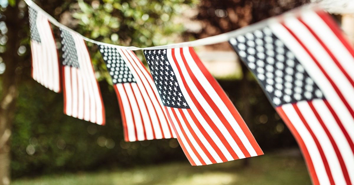A row of small American flags is strung together, hanging outdoors in a sunlit garden or park setting. The flags are gently swaying, with a blurred green background suggesting trees and foliage. The image evokes a festive, patriotic mood, ideal for national celebrations or holidays.
