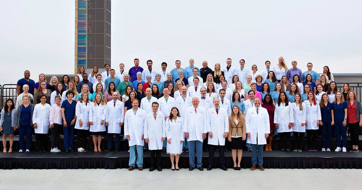 A large group of people, including men and women of various ages, are posed together outdoors on a raised platform. Many individuals in the front row are wearing white lab coats, while others are dressed in business or casual attire. The background features a tall building and an overcast sky, suggesting an institutional or academic setting. The group appears to be a team of healthcare or medical staff gathered for a formal group photo.