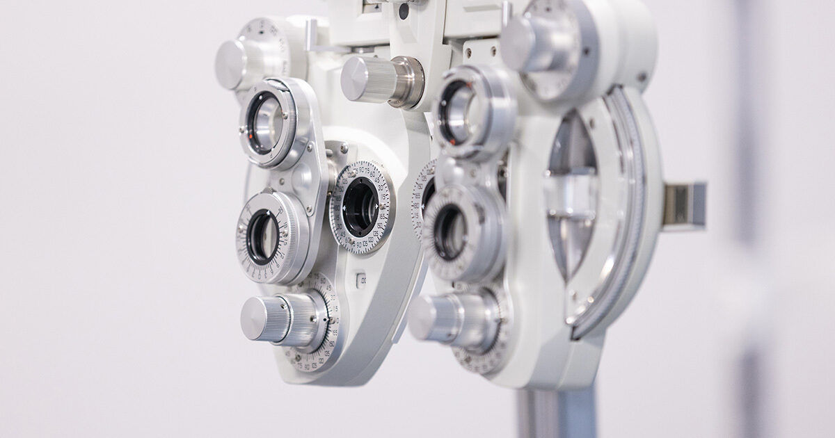 A detailed view of a phoropter, an essential device used in eye examinations to measure refractive error and determine eyeglass prescriptions. The equipment is shown in a clinical setting with a clean, neutral background, highlighting its metallic dials and lenses. The image focuses on the intricate mechanical components, emphasizing precision and advanced technology. No visible text or numbers are present on the device.