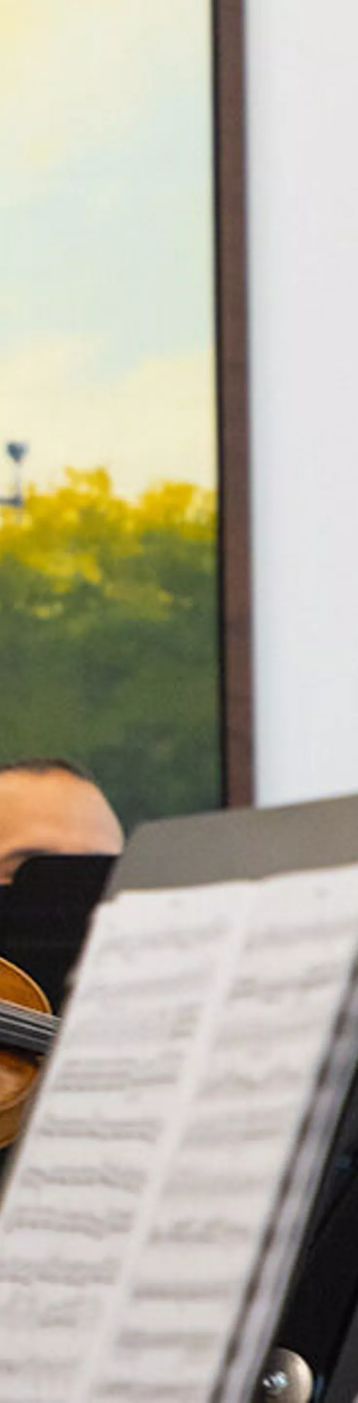 A group of musicians is performing together, each playing a violin in an indoor setting. The background features a large framed painting of a cityscape with a prominent dome structure and lampposts. Sheet music is visible in the foreground, indicating a formal or practice session. The overall mood is focused and collaborative, with natural daylight illuminating the scene.