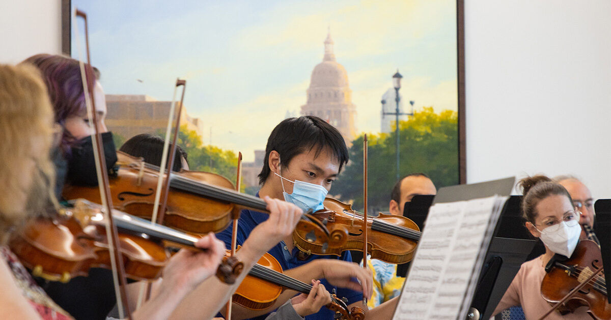 A group of musicians is performing together, each playing a violin in an indoor setting. The background features a large framed painting of a cityscape with a prominent dome structure and lampposts. Sheet music is visible in the foreground, indicating a formal or practice session. The overall mood is focused and collaborative, with natural daylight illuminating the scene.