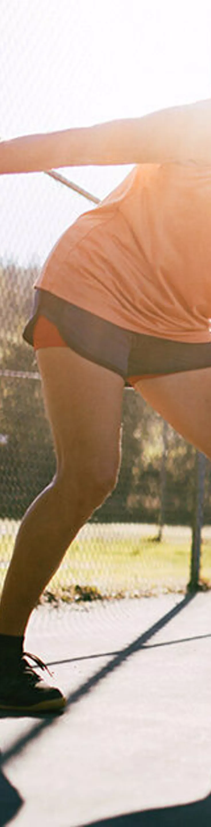 Two adults are actively engaged in a game of pickleball on an outdoor court under bright sunlight. Both players hold paddles and are positioned to return the ball, with a chain-link fence and trees visible in the background. The scene captures dynamic movement and a casual, energetic atmosphere. The lighting is warm, suggesting a sunny day. No visible text or numbers are present in the image.