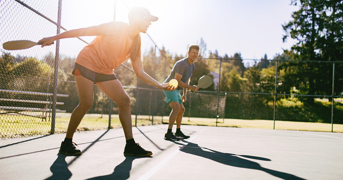 Two adults are actively engaged in a game of pickleball on an outdoor court under bright sunlight. Both players hold paddles and are positioned to return the ball, with a chain-link fence and trees visible in the background. The scene captures dynamic movement and a casual, energetic atmosphere. The lighting is warm, suggesting a sunny day. No visible text or numbers are present in the image.