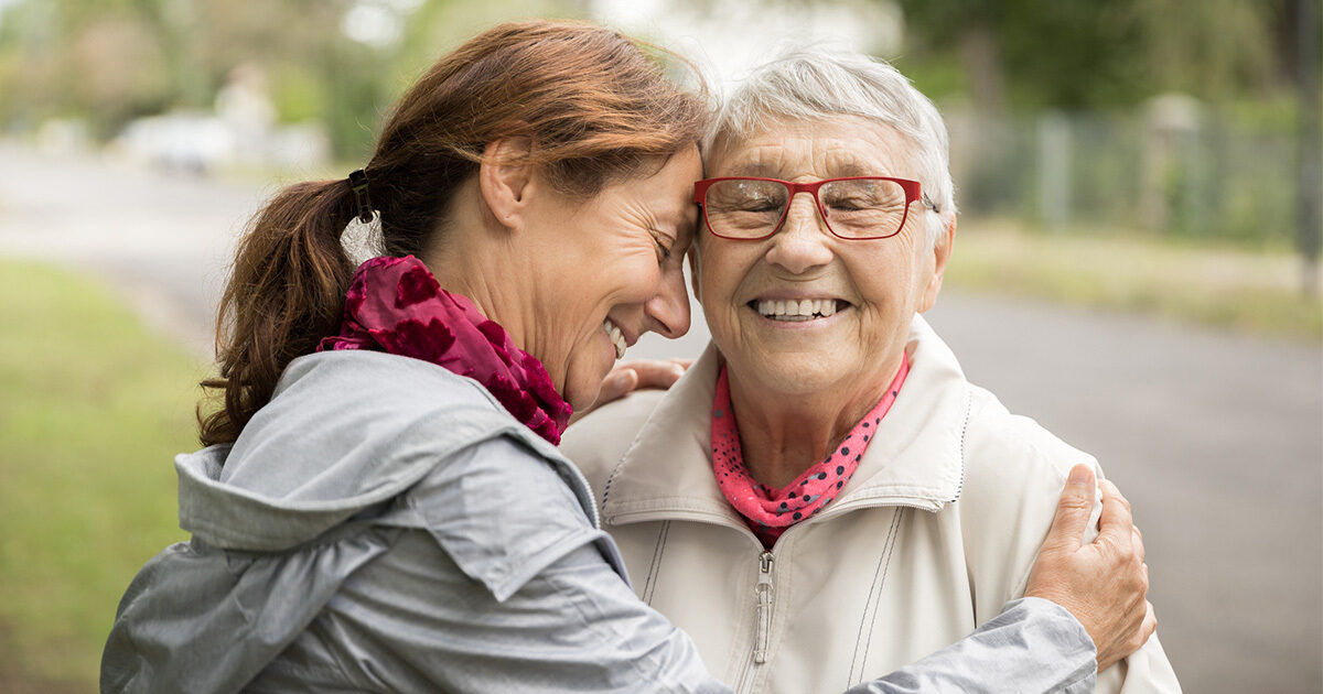 Two women are seen embracing each other outdoors, suggesting a moment of comfort or support. Both are dressed in light jackets, with one wearing a grey jacket and the other in a cream jacket, each accessorized with a pink scarf. The background features a blurred outdoor setting with greenery and a pathway, indicating a casual or supportive interaction in a park or residential area. The overall mood is caring and gentle.
