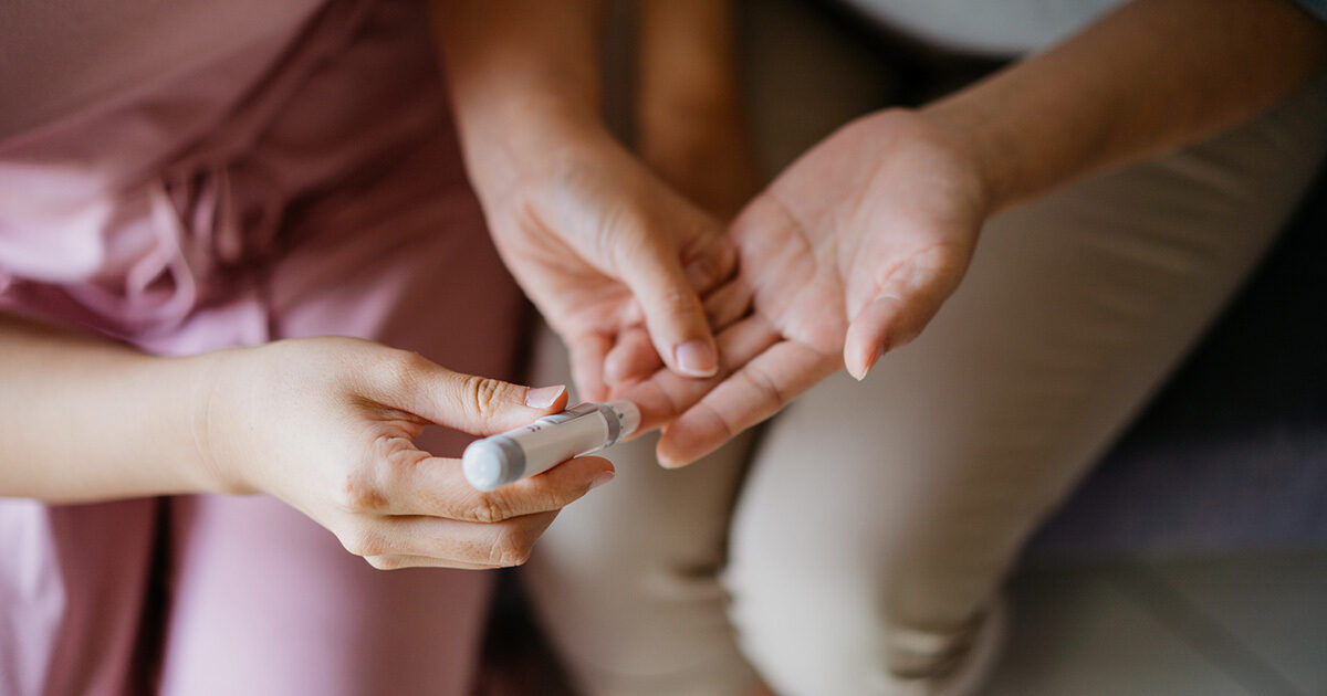 A close-up view shows a woman preparing to check another person's blood sugar using a lancet device. The setting appears to be indoors, possibly at home, with soft natural lighting. The focus is on the hands and the medical device, emphasizing a moment of care and health monitoring. The image conveys a calm and supportive atmosphere.