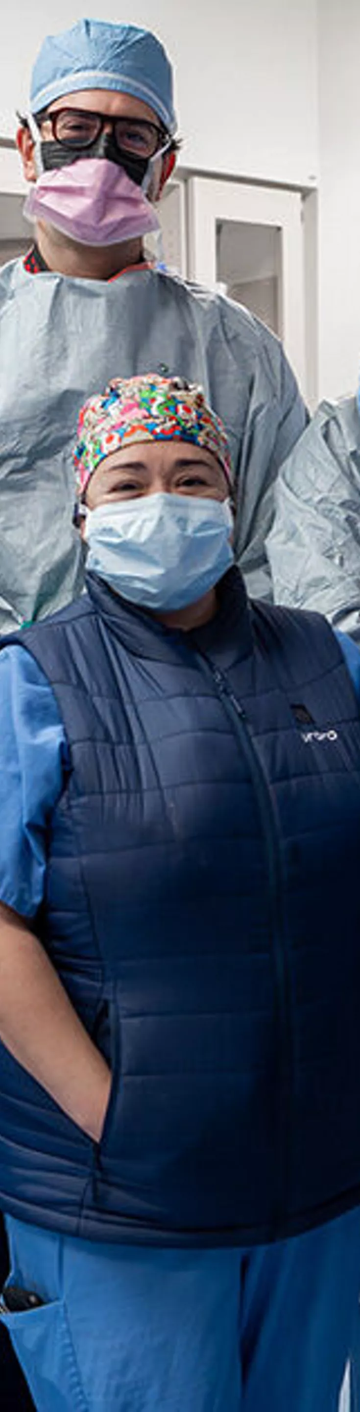 A group of healthcare professionals stands together in a hospital operating room, dressed in full surgical attire including gowns, gloves, and caps. The setting features medical equipment, monitors, and cabinets in the background, indicating a sterile surgical environment. The team appears to be preparing for or concluding a medical procedure. The overall mood is professional and collaborative, with a clinical blue and white color palette.