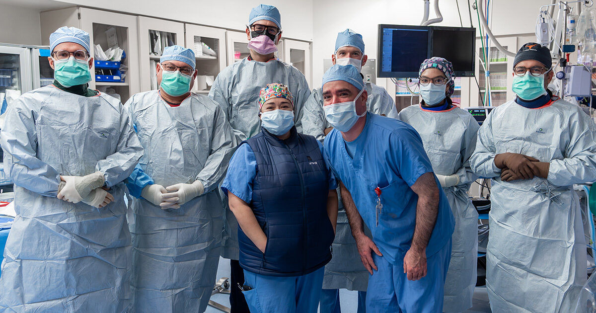 A group of healthcare professionals stands together in a hospital operating room, dressed in full surgical attire including gowns, gloves, and caps. The setting features medical equipment, monitors, and cabinets in the background, indicating a sterile surgical environment. The team appears to be preparing for or concluding a medical procedure. The overall mood is professional and collaborative, with a clinical blue and white color palette.