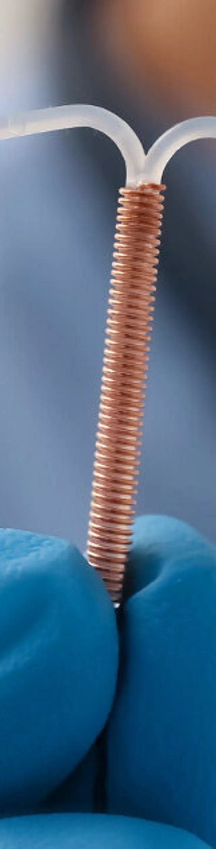 A gloved hand holds a copper intrauterine device (IUD) in focus, with a healthcare professional in blue scrubs visible in the background. The setting appears to be a clinical or medical environment, emphasizing a sterile and professional atmosphere. The IUD's copper coil and T-shape are clearly visible, highlighting its use in contraception. The image conveys a sense of trust and medical expertise.