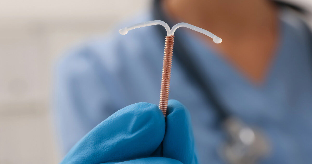 A gloved hand holds a copper intrauterine device (IUD) in focus, with a healthcare professional in blue scrubs visible in the background. The setting appears to be a clinical or medical environment, emphasizing a sterile and professional atmosphere. The IUD's copper coil and T-shape are clearly visible, highlighting its use in contraception. The image conveys a sense of trust and medical expertise.