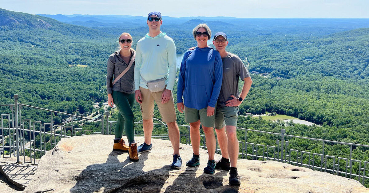 Four people stand together on a rocky mountain overlook, surrounded by expansive green forest and distant hills under a clear blue sky. The group is dressed in casual outdoor attire, including shorts, athletic shoes, and a crossbody bag. A metal safety railing lines the edge of the viewpoint, emphasizing the elevated location. Bright sunlight and vivid colors create a cheerful, adventurous mood. No visible text or numbers are present in the image.