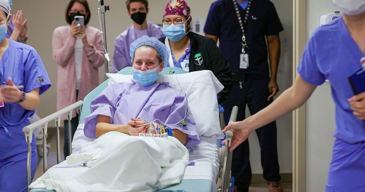 A patient lies on a hospital bed, covered with a white blanket and connected to monitoring equipment. Several healthcare professionals in scrubs and uniforms accompany the patient through a clinical hallway. The setting is a modern hospital, with a focus on medical care and teamwork. The scene conveys a sense of urgency and professionalism, with muted colors and clinical lighting. No visible text or numbers are present in the image.
