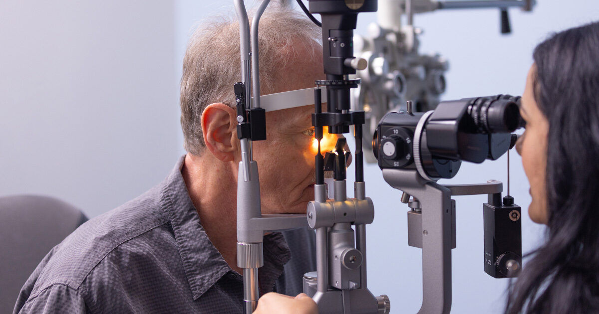 An adult male patient undergoes an eye examination using a slit lamp, a specialized ophthalmic instrument. The setting is a clinical optometry office with medical equipment visible in the background. A female optometrist or technician operates the device, focusing on the patient's eye health. The scene is well-lit with neutral tones, emphasizing a professional healthcare environment.