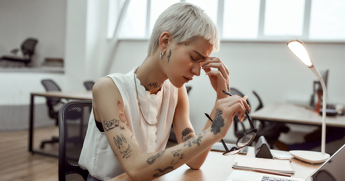 A woman with short platinum blonde hair and visible tattoos sits at a contemporary office desk. She is holding eyeglasses and appears to be focused on her work, surrounded by a laptop, documents, and a desk lamp. The setting is a bright, open workspace with large windows and minimalist decor.