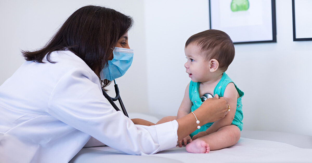 A healthcare professional uses a stethoscope to examine a baby sitting on an examination table. The setting is a bright, clean medical office with framed artwork on the wall. The baby is wearing a turquoise outfit and appears calm during the checkup. The scene conveys a caring and professional healthcare environment.