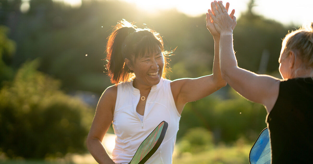 Two women are seen playing pickleball outdoors, sharing a high-five in celebration. The setting is a sunlit park or recreational area, with lush greenery in the background. Both women hold pickleball paddles, and the scene conveys a joyful, active mood. The lighting is warm and golden, suggesting late afternoon or early evening.