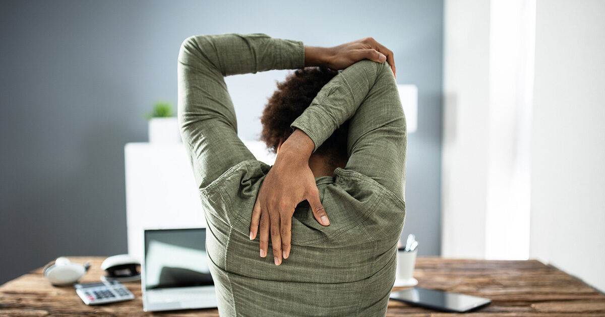 An adult is seen stretching their arms behind their head while seated at a wooden desk in a modern office setting. The workspace includes a laptop, calculator, and documents, suggesting a work-from-home or office environment. The person is viewed from behind, emphasizing relaxation or a break during work. The overall mood is calm and focused, with neutral and natural tones dominating the scene.