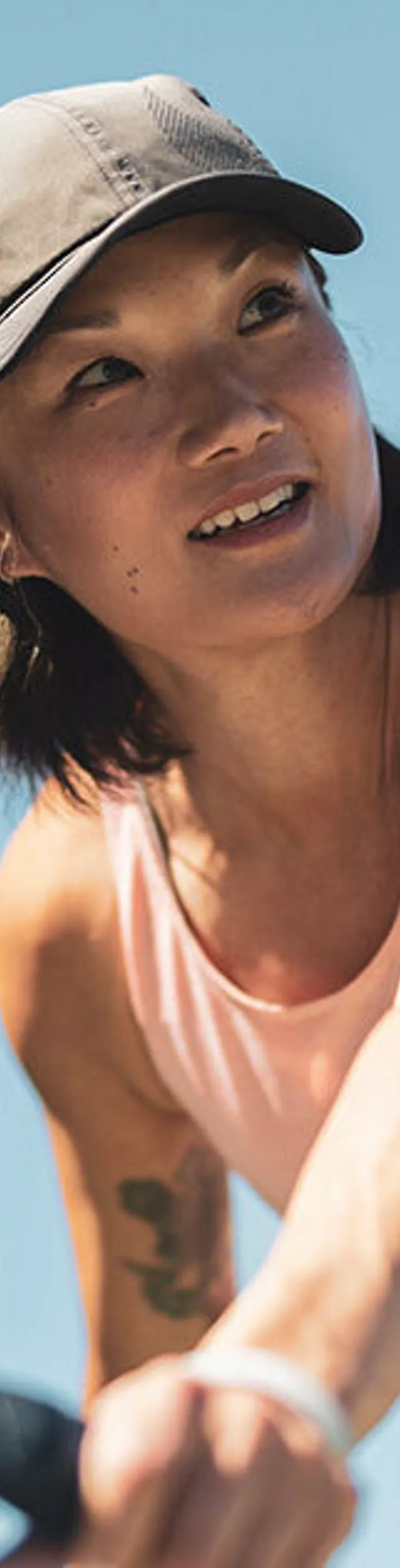 An adult woman is actively playing pickleball outdoors, captured mid-action as she prepares to hit a yellow ball with a black paddle. She is wearing a light pink sleeveless top and a cap, with a clear blue sky in the background. The image conveys energy and focus, with bright natural lighting and a sporty atmosphere.