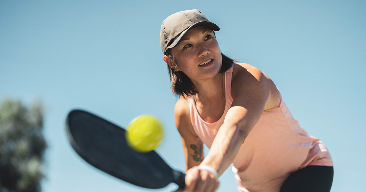 An adult woman is actively playing pickleball outdoors, captured mid-action as she prepares to hit a yellow ball with a black paddle. She is wearing a light pink sleeveless top and a cap, with a clear blue sky in the background. The image conveys energy and focus, with bright natural lighting and a sporty atmosphere.