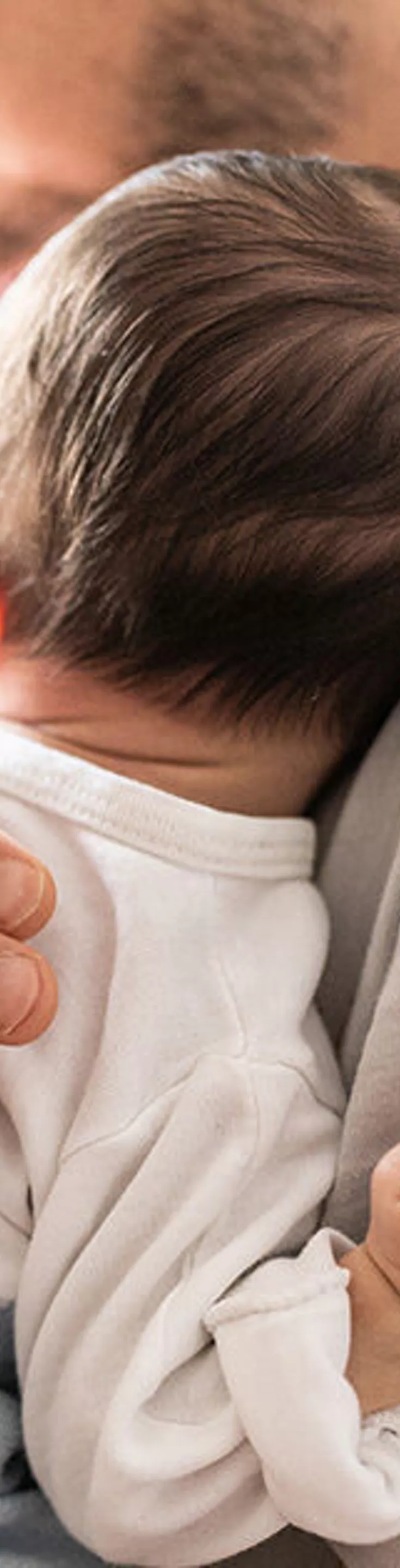 An adult gently holds a newborn baby against their chest in a softly lit room. The scene is set near a window with sheer curtains, allowing natural light to fill the space. The baby is dressed in a white and blue outfit, and the overall mood is calm and nurturing. The focus is on the tender interaction and the peaceful environment.