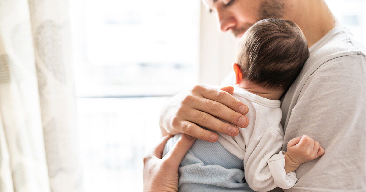 An adult gently holds a newborn baby against their chest in a softly lit room. The scene is set near a window with sheer curtains, allowing natural light to fill the space. The baby is dressed in a white and blue outfit, and the overall mood is calm and nurturing. The focus is on the tender interaction and the peaceful environment.