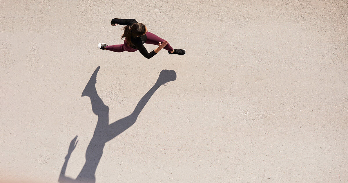 An overhead view shows a woman in athletic wear running on a light concrete surface. Her dynamic pose and extended shadow create a striking visual effect. The setting is minimalistic, emphasizing movement and energy. The image captures a sense of motion and determination.