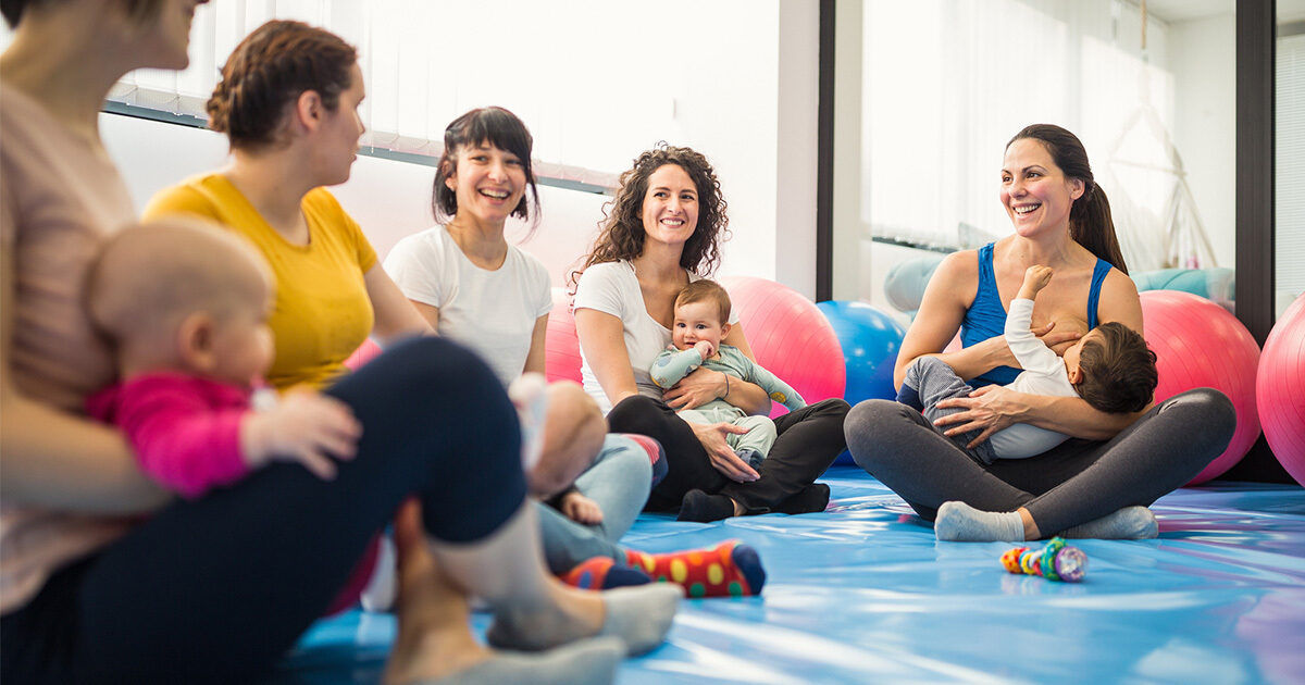 A group of women with infants are seated on a blue mat in a bright, spacious room. Large exercise balls are visible in the background, suggesting a fitness or parenting class setting. The women are engaged in conversation and caring for their babies, creating a supportive and relaxed atmosphere. The scene features casual attire and soft, natural lighting.