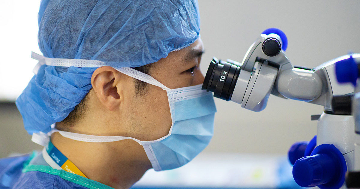 A healthcare worker wearing a blue surgical cap and mask is closely examining something through a laboratory microscope. The setting appears to be a medical or research environment, with the individual focused on the optical device. The image highlights the use of advanced technology in healthcare and scientific research. The overall palette is clean and clinical, dominated by blue and white tones.