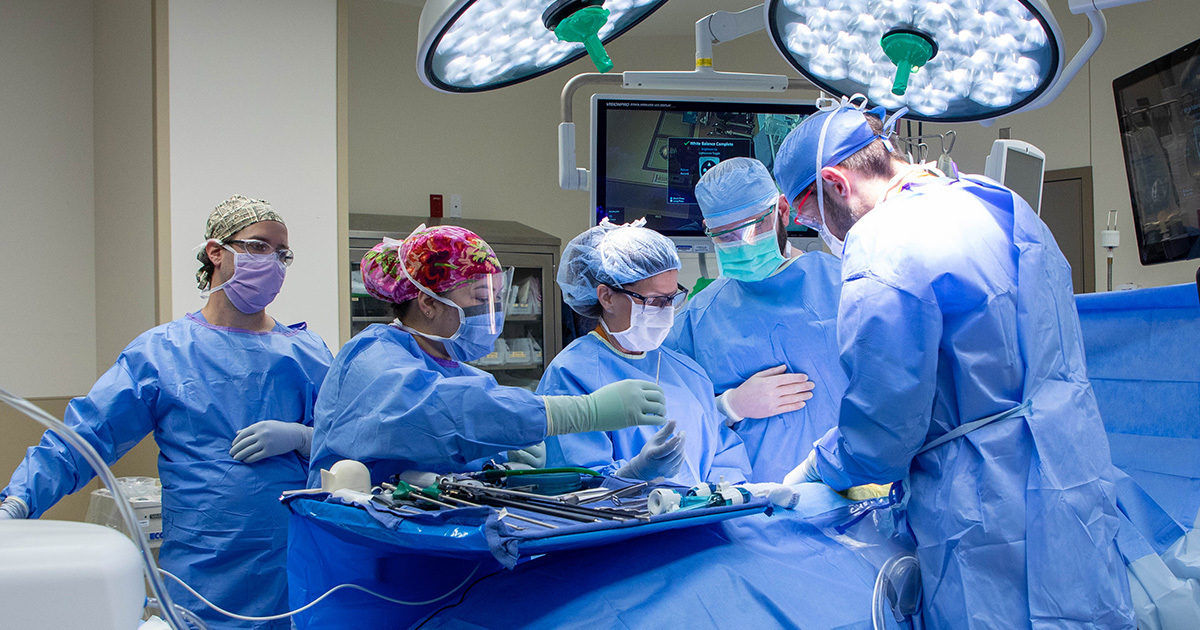 A group of medical professionals is engaged in a surgical procedure under bright operating lights. The setting is a sterile hospital operating room, with surgical instruments and monitors visible. The team is dressed in blue surgical gowns, masks, and caps, focusing intently on the patient. The environment is clean and highly organized, emphasizing precision and teamwork.