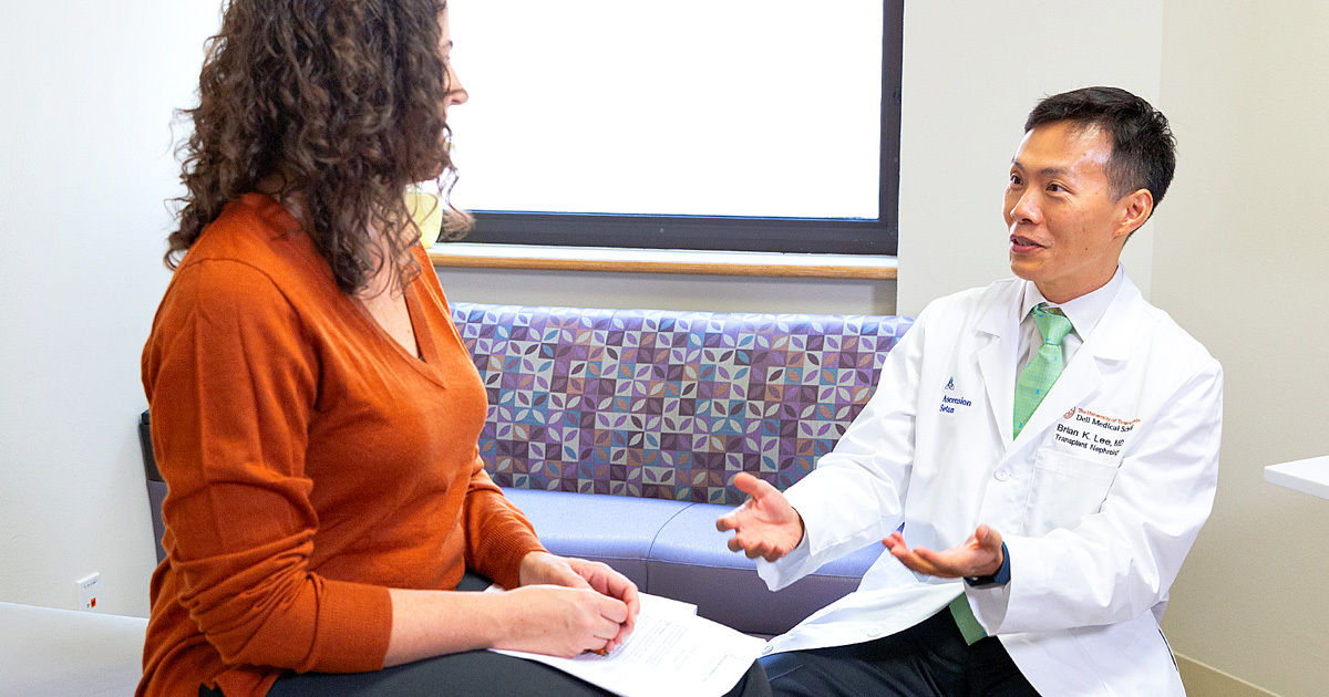 A healthcare professional in a white coat speaks with a seated woman in an orange sweater. The setting is a modern medical office with natural light and patterned seating. The doctor gestures with open hands, suggesting a discussion or consultation. The environment is clean and professional, with a calm and approachable atmosphere.
