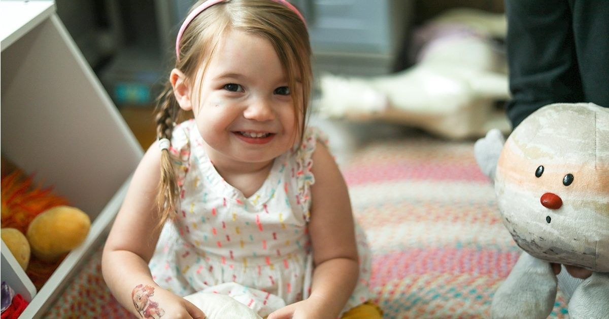 A young girl sits on a colorful patterned rug in a cozy bedroom, holding a soft toy. She is dressed in a light-colored dress with small patterns and a pink headband. The background features plush toys and soft furnishings, creating a warm and playful atmosphere. The scene suggests a relaxed, indoor playtime setting.