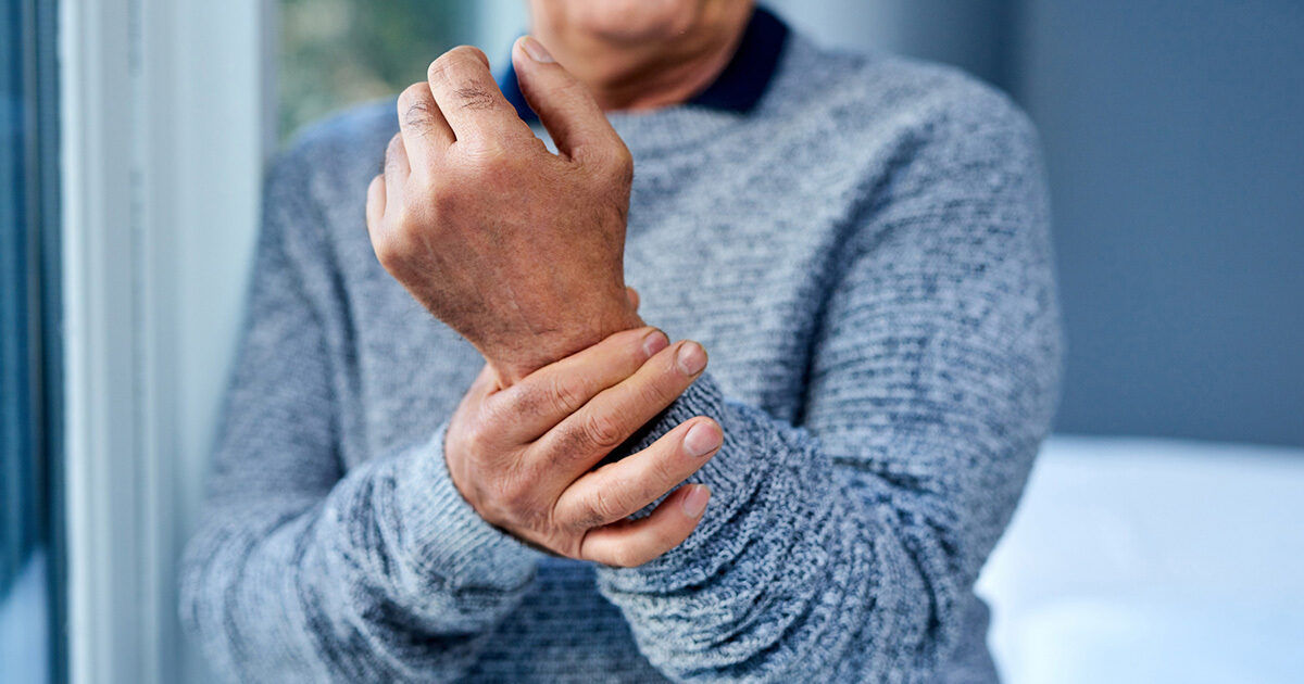An adult man is shown holding his wrist, suggesting discomfort or pain. He is dressed in a grey sweater and is indoors, likely in a home environment. The image focuses on his hands and torso, with a soft, neutral background. No visible text or numbers are present.
