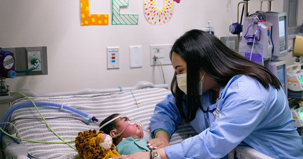 A nurse leans over an infant lying in a hospital crib, providing comfort and care. The room contains medical equipment and monitors, with colorful letters spelling 'LEO' on the wall above the crib. The infant is accompanied by a plush toy, and various tubes and wires are visible, indicating a medical environment. The scene conveys a caring and supportive atmosphere.