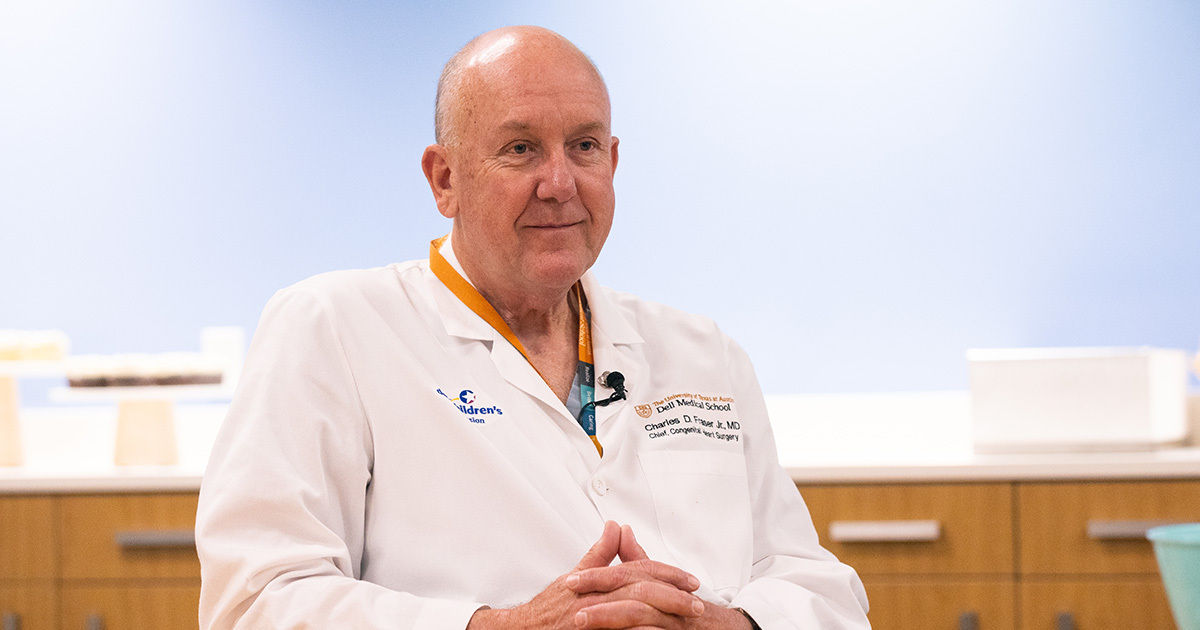 A medical professional is seated in a clinical setting, wearing a white lab coat with visible embroidered text and a stethoscope. The background features a light blue wall and wooden cabinetry, suggesting a modern healthcare environment. The doctor's hands are folded, and the overall atmosphere is clean and professional. Some text is visible on the coat, including a name and medical credentials.