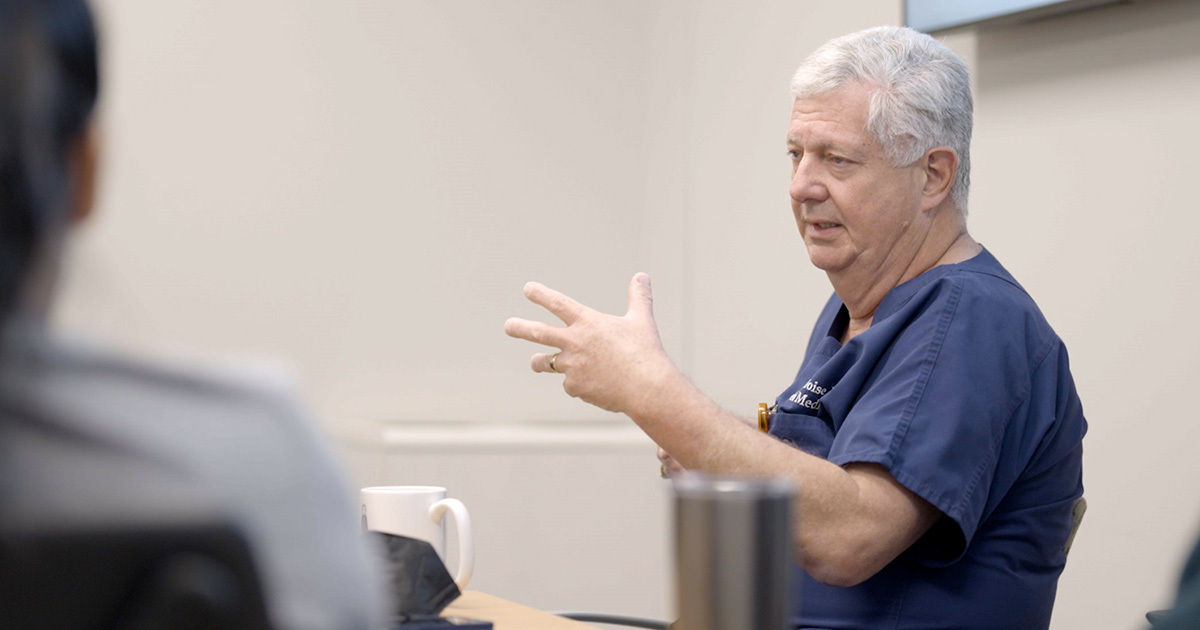 An older adult in medical scrubs gestures while speaking during a meeting in a clinical office setting. The environment includes a table, a coffee mug, and a stainless steel tumbler, suggesting a collaborative discussion. The focus is on the person's expressive hand movements and professional attire. The background is neutral, emphasizing a calm and professional atmosphere.