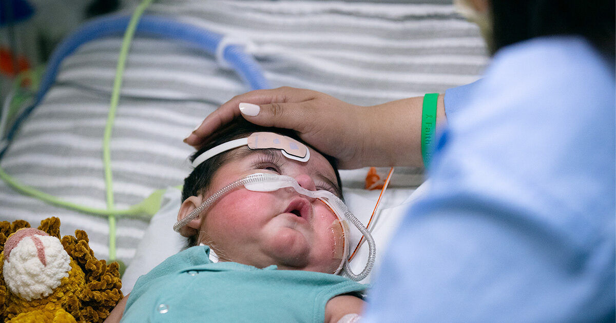 An adult gently rests a hand on an infant lying in a hospital bed. The baby is wearing a medical gown and has tubes attached, indicating a medical procedure or care. A soft toy is visible beside the infant, adding a comforting touch. The environment suggests a clinical or pediatric care setting, with striped bedding and medical equipment present.