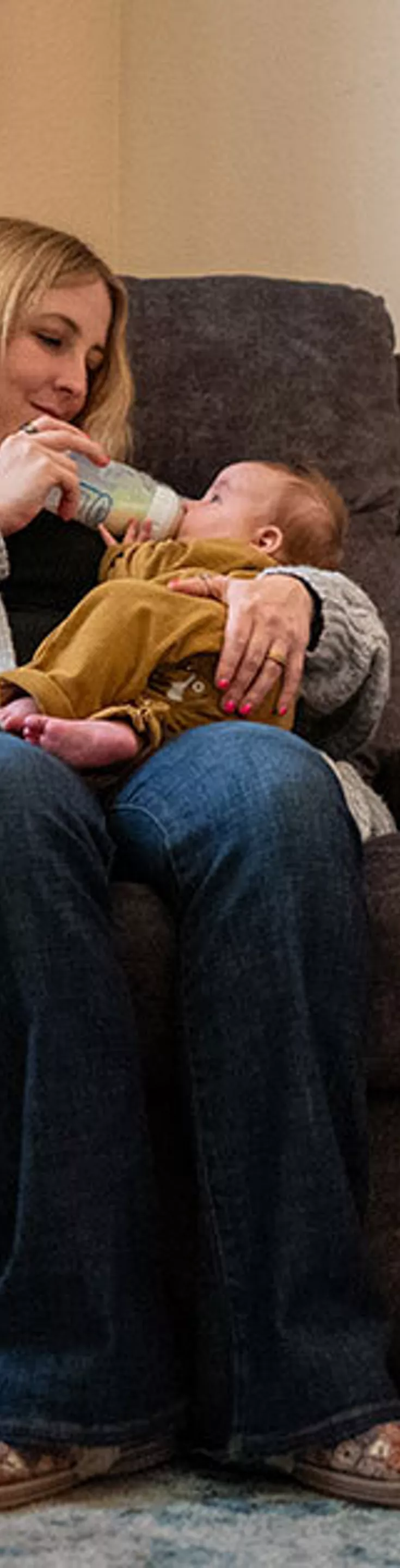 A woman sits in a plush armchair feeding a baby with a bottle in a softly lit nursery. The room features a changing table stocked with diapers, wipes, and baby essentials, as well as a side table with lotion and sanitizer. The setting is warm and homey, with muted colors and gentle lighting creating a calm atmosphere. Visible items include baby care products and organized storage bins.
