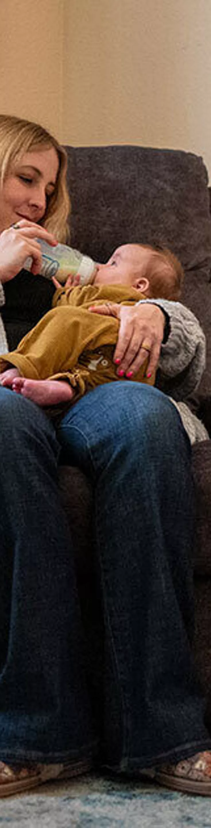A woman sits in a plush armchair feeding a baby with a bottle in a softly lit nursery. The room features a changing table stocked with diapers, wipes, and baby care items, as well as a small side table with baby essentials. The environment is warm and inviting, with neutral tones and soft textures. Visible baby products and organized supplies highlight a nurturing atmosphere.