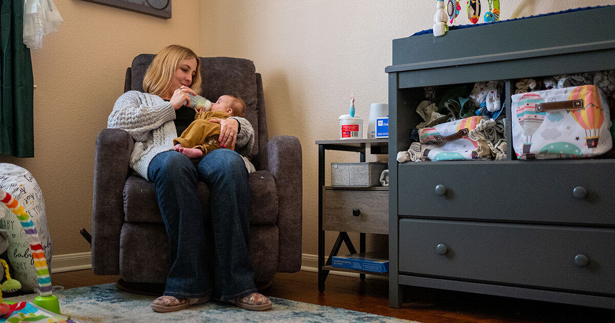 A woman sits in a plush armchair feeding a baby with a bottle in a softly lit nursery. The room features a changing table stocked with diapers, wipes, and baby care items, as well as a small side table with baby essentials. The environment is warm and inviting, with neutral tones and soft textures. Visible baby products and organized supplies highlight a nurturing atmosphere.