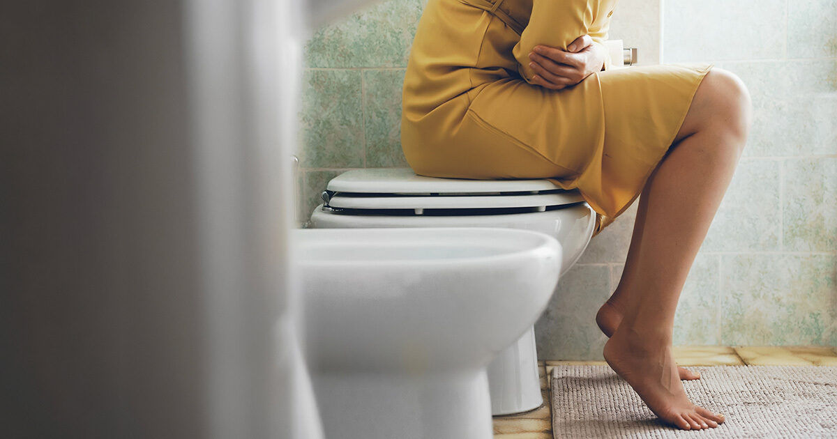 A woman wearing a yellow dress is seated on a closed toilet in a tiled bathroom. She appears to be holding her stomach, possibly indicating discomfort or pain. The setting includes a white toilet, tiled walls, and a bath mat on the floor. The image conveys a private, everyday moment with a neutral to somber mood. No visible text or numbers are present.