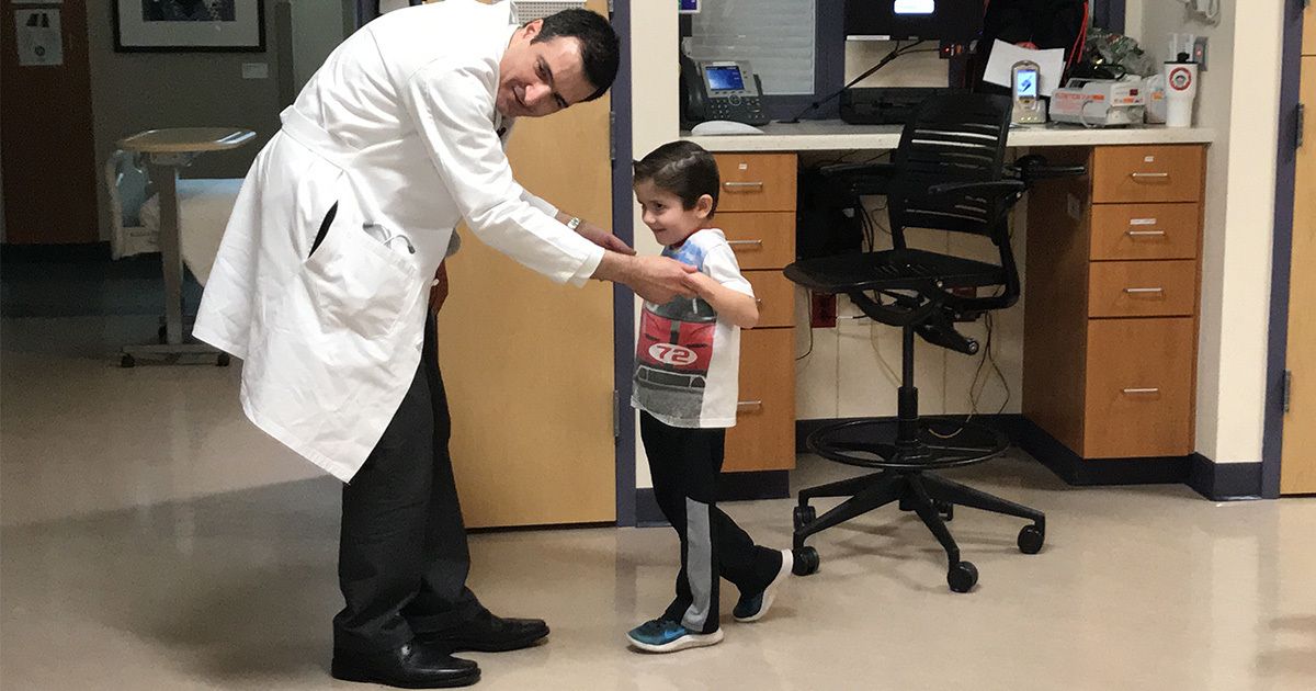 A male doctor in a white coat helps a young boy walk in a hospital or clinical setting. The child is wearing a prosthetic leg and casual clothing, while the doctor gently supports him. The background shows medical equipment, a desk, and cabinets, indicating a healthcare environment. The scene conveys care, support, and rehabilitation.