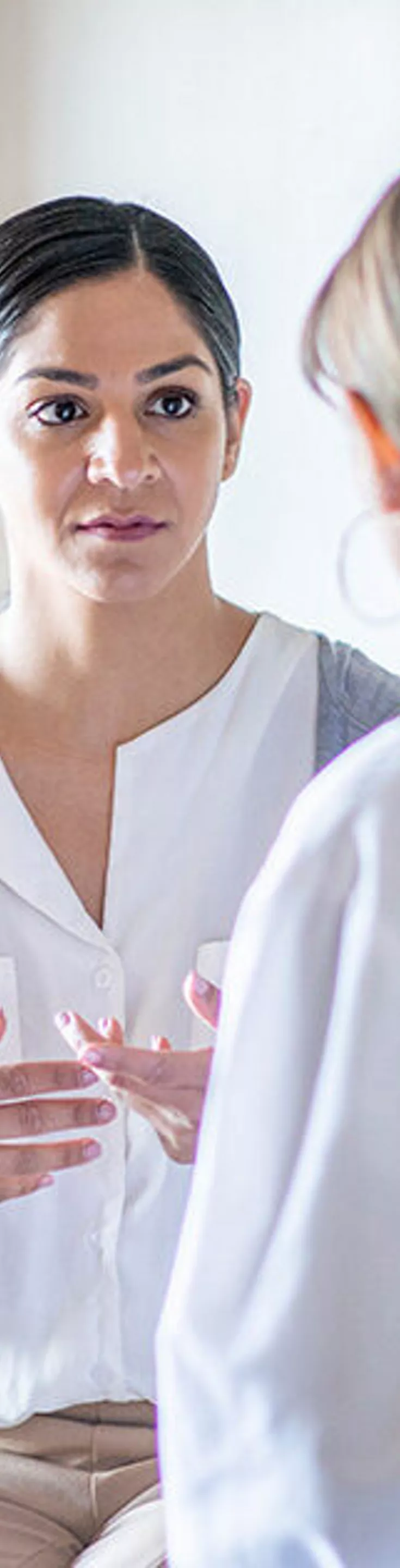 A patient sits on an examination table while speaking with a healthcare professional in a clinical setting. The room is well-lit with natural light coming from a window. Medical equipment is visible on the wall, and both individuals appear engaged in conversation. The scene conveys a calm and professional atmosphere.